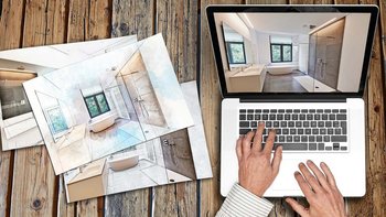 Auf einem Holztisch liegen Skizzen eines modernen Badezimmers, während eine Person am Laptop arbeitet.