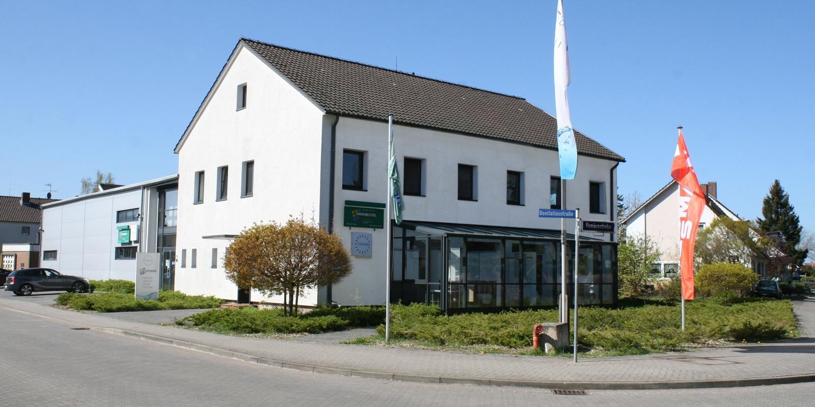 Die Fassade des Gebäudes zeigt große Fenster und ein Schild mit dem Namen 'Hornbostel', während bunte Fahnen im Wind wehen.